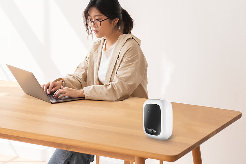 A woman working on a laptop at a wooden table, with a Shizen SBX1 on the table beside her