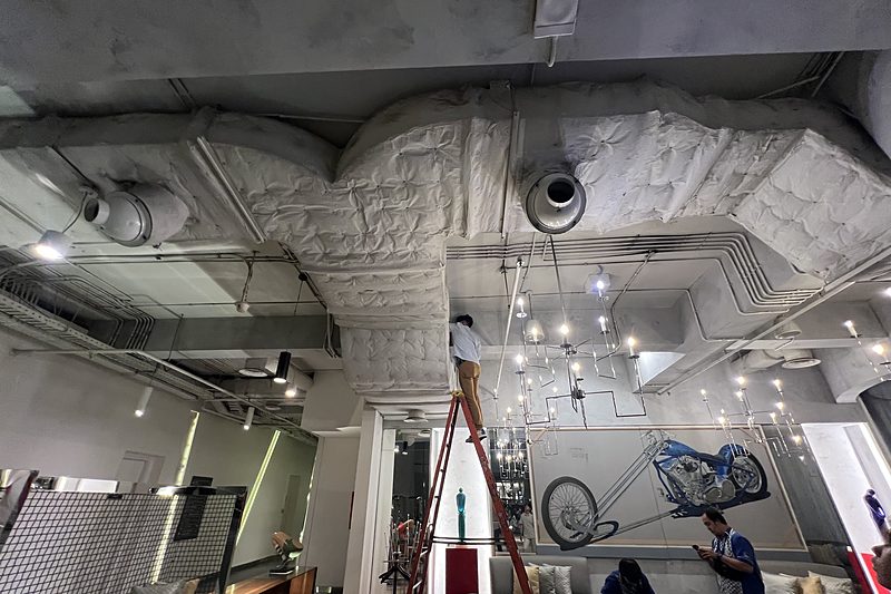 A technician on a stepladder working on insulated air-conditioning ducts in the ceiling of a hotel lounge, with two colleagues below