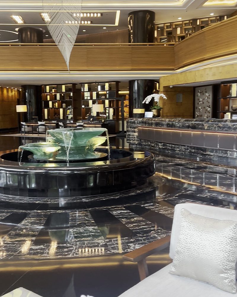 A hotel lobby with a dark marble floor, a fountain, the reception desk and a white sofa
