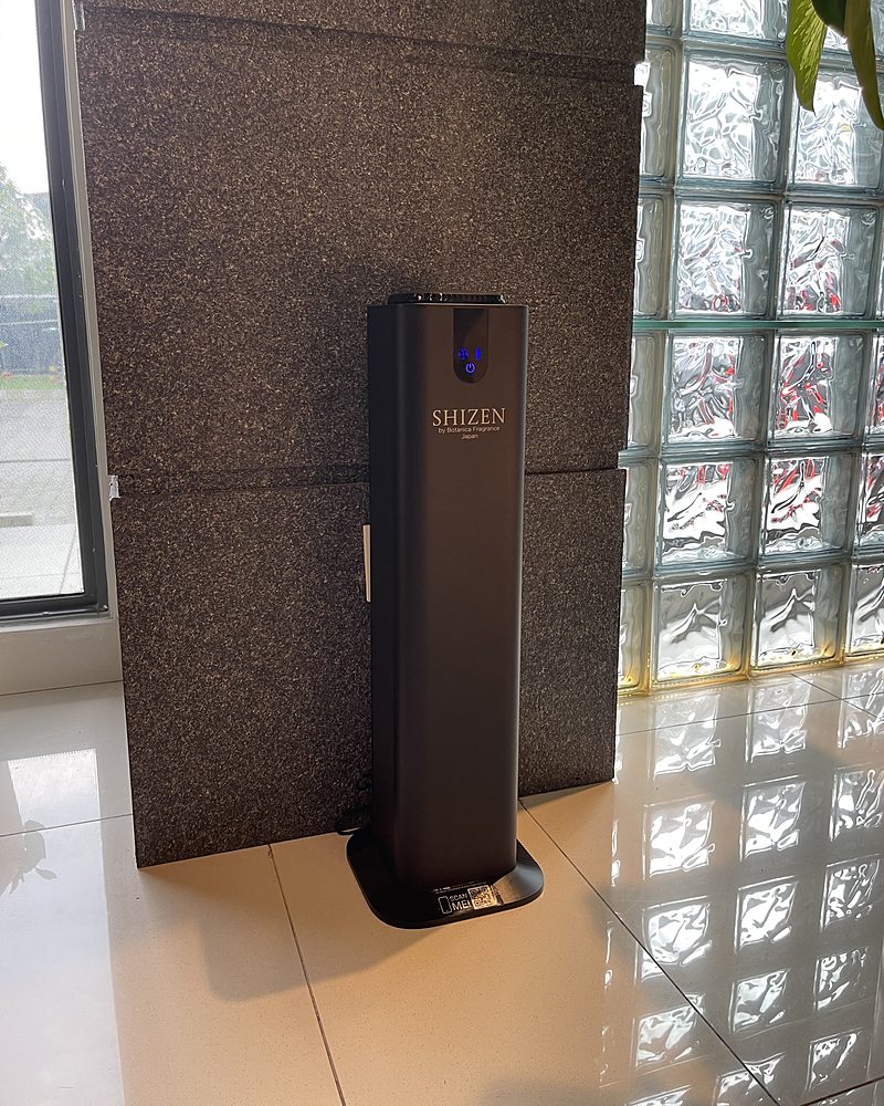 A black tower-shaped Shizen SBA2 scent diffuser in an office lobby beside a granite pillar and a glass-block wall