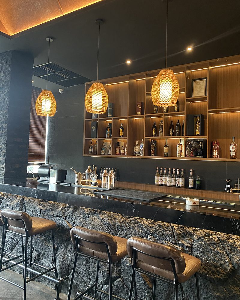 A stone bar counter with leather stools, lit bottle shelves and rattan pendant lamps