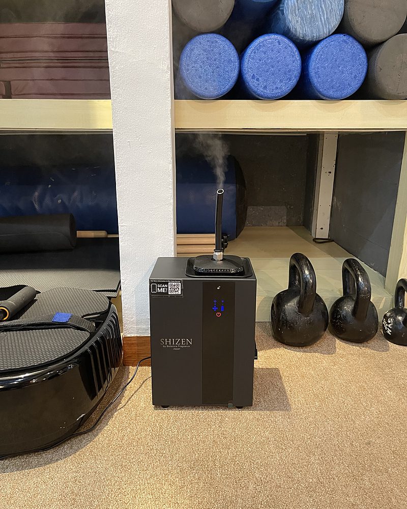 A black Shizen SBA1 releasing a thin mist from the nozzle on top, beside kettlebells and foam rollers in a studio